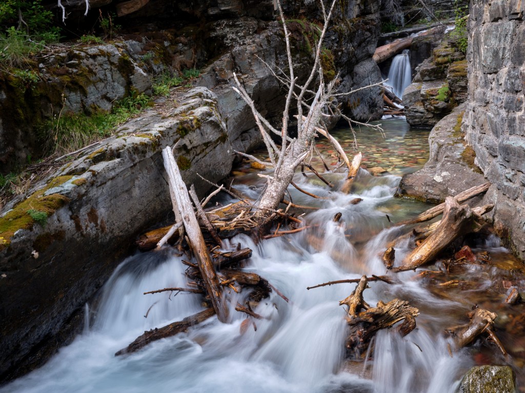 A mountain creek rushes through a narrow channel filled with driftwood, stones, and blurred whitewater.