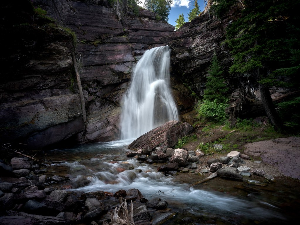 Silky white water spills from a cliff into a shallow stream bordered by dark stone and forest.