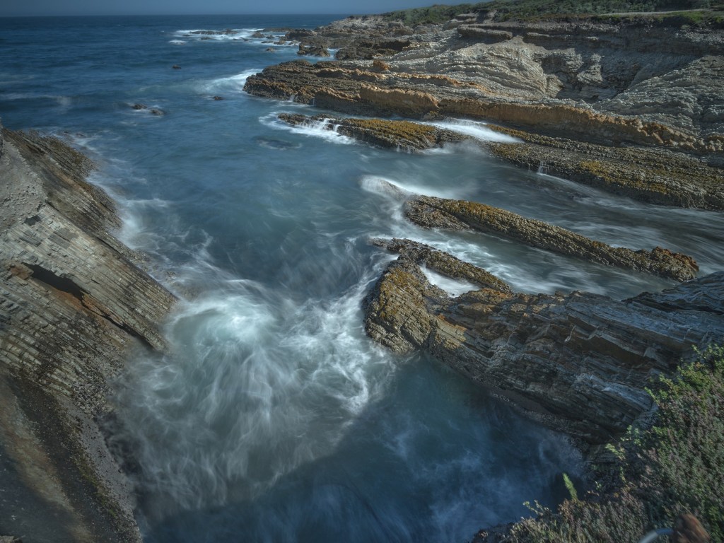A high coastal view shows swirling currents between tan formations and dark ocean channels.