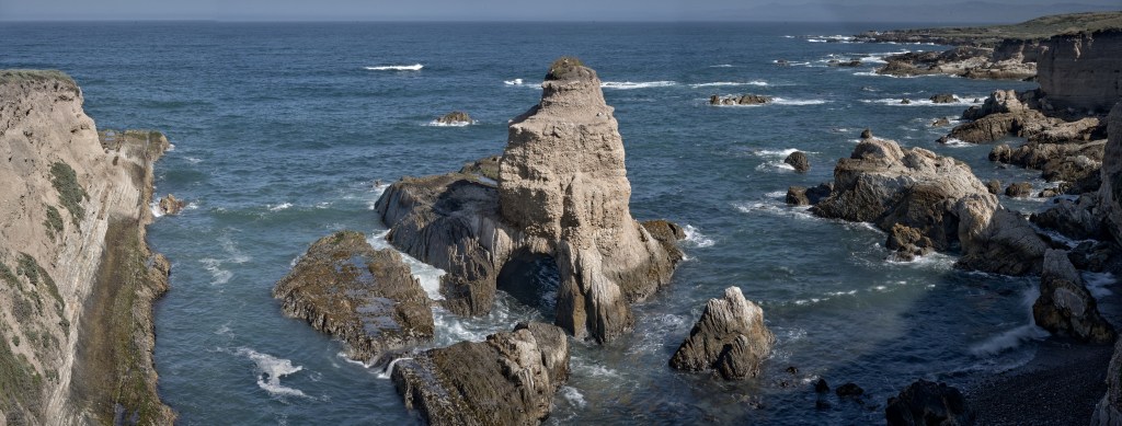 Wide coastal view with eroded rock towers, blue ocean, and layered headlands along the trail.