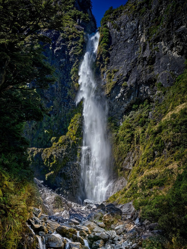 New Zealand 42 A high white waterfall drops between dark rock walls surrounded by dense green mountain forest.