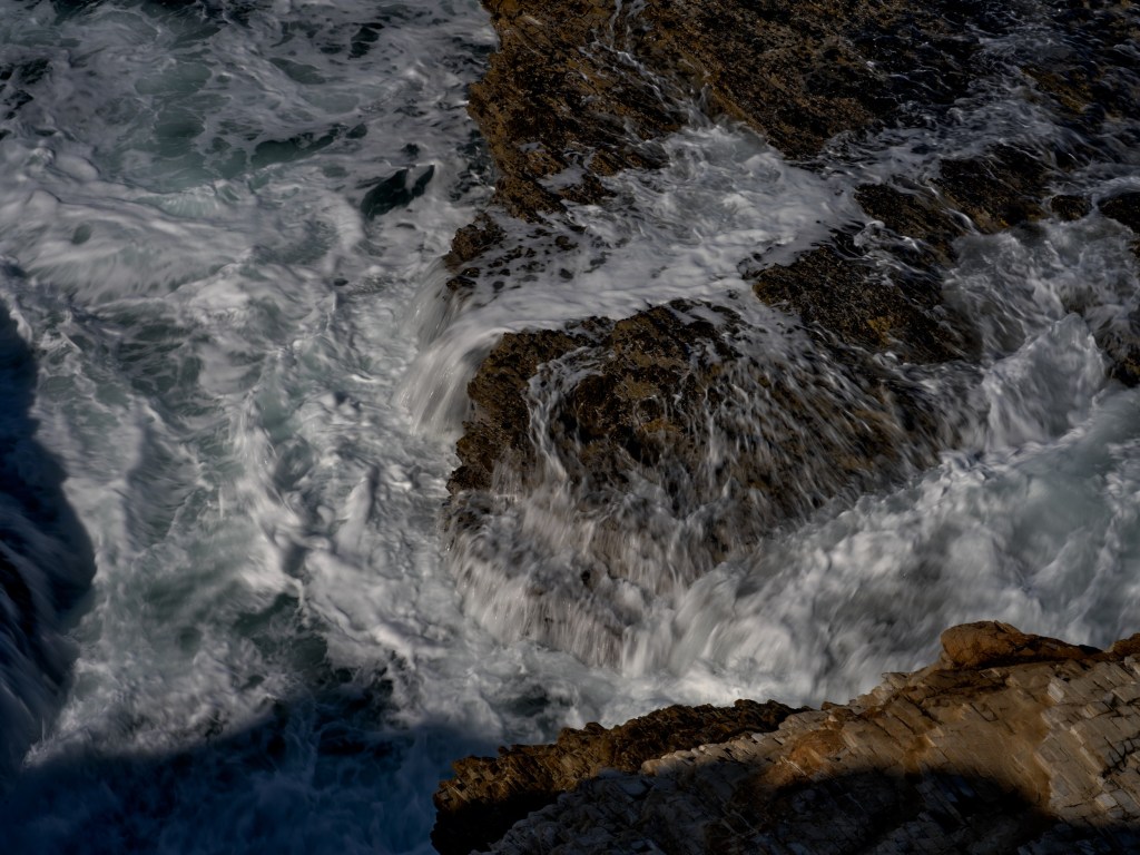 A high viewpoint captures abstract whitewater patterns wrapping around submerged shoreline stone.