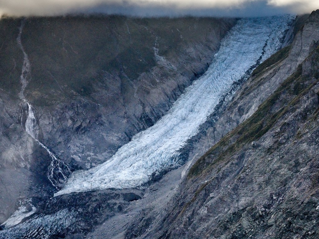 New Zealand 58 A pale glacier tongue snakes down a steep valley with waterfalls and cloud shadows nearby.
