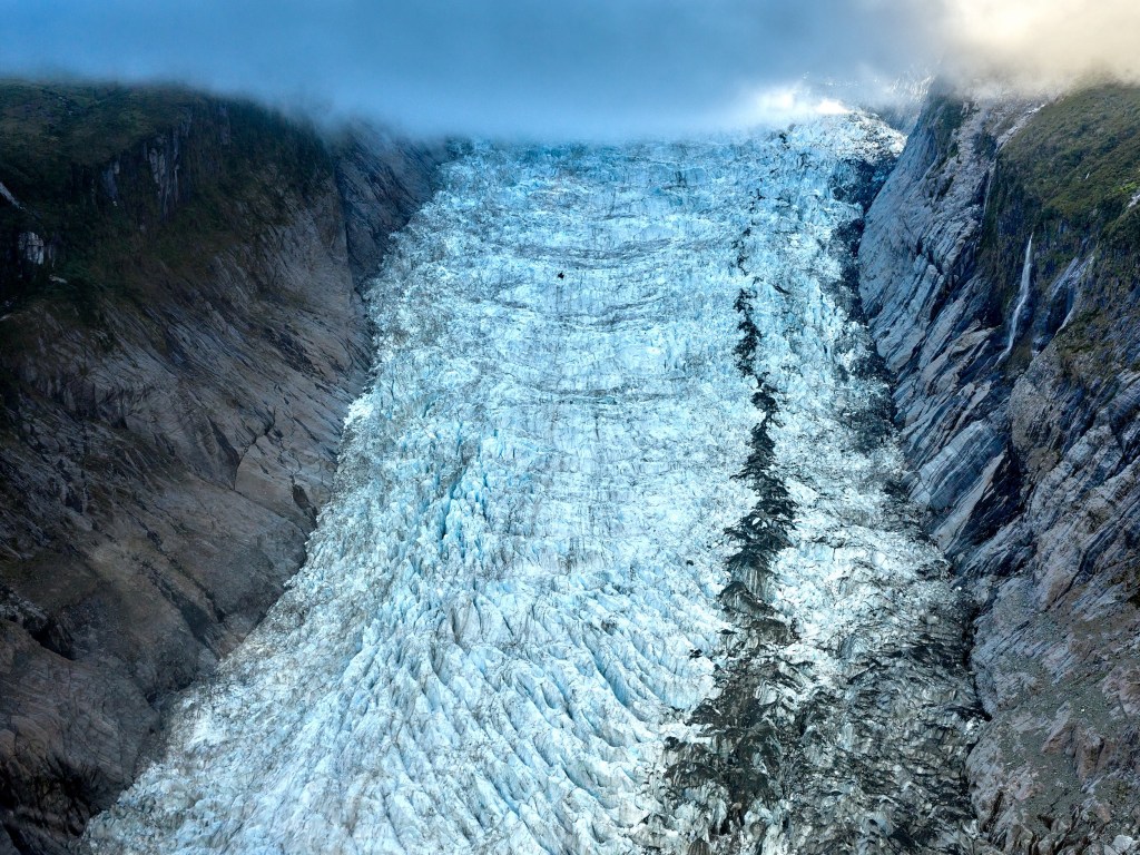New Zealand 11 Blue white glacier ice fills a narrow valley and fades upward into mist and cloud.