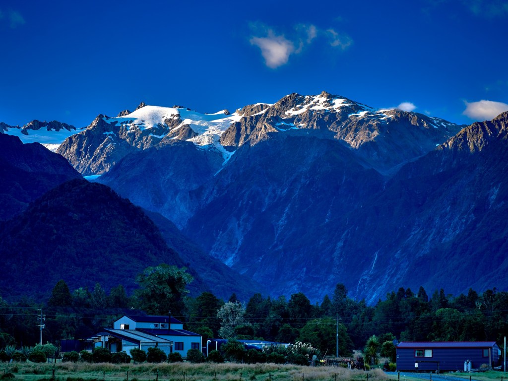 New Zealand 39 Early sunlight catches snow covered summits above the dark village and forested valley.