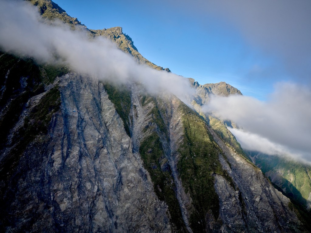 New Zealand 68 Steep gray cliffs and passing cloud reveal the rugged terrain below Franz Josef Glacier.