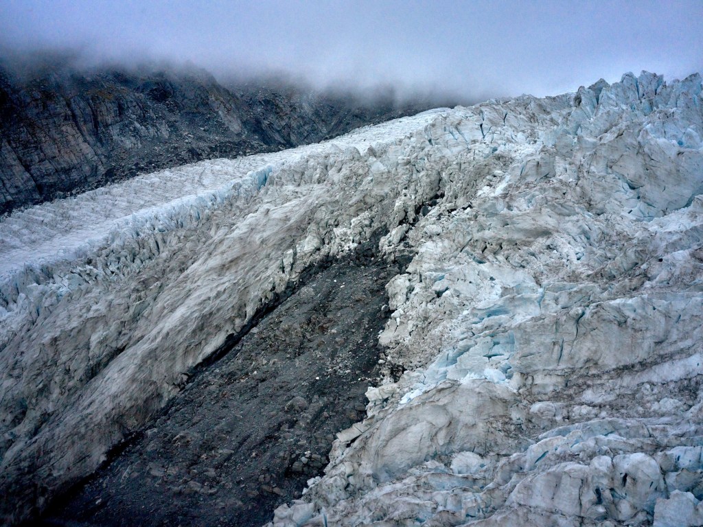 New Zealand 10 Crevassed glacier ice flows down a dark valley toward rocky slopes and misty cloud.