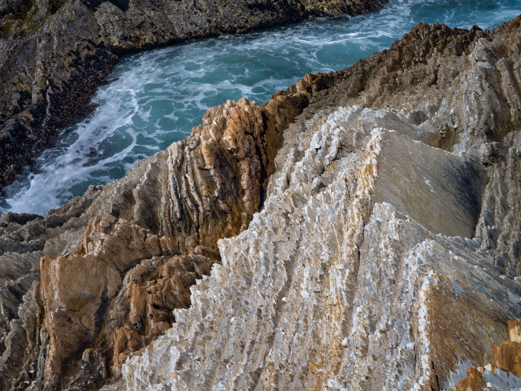 Steep folded sandstone ridges descend toward a narrow pocket of surf along the rugged coastline.