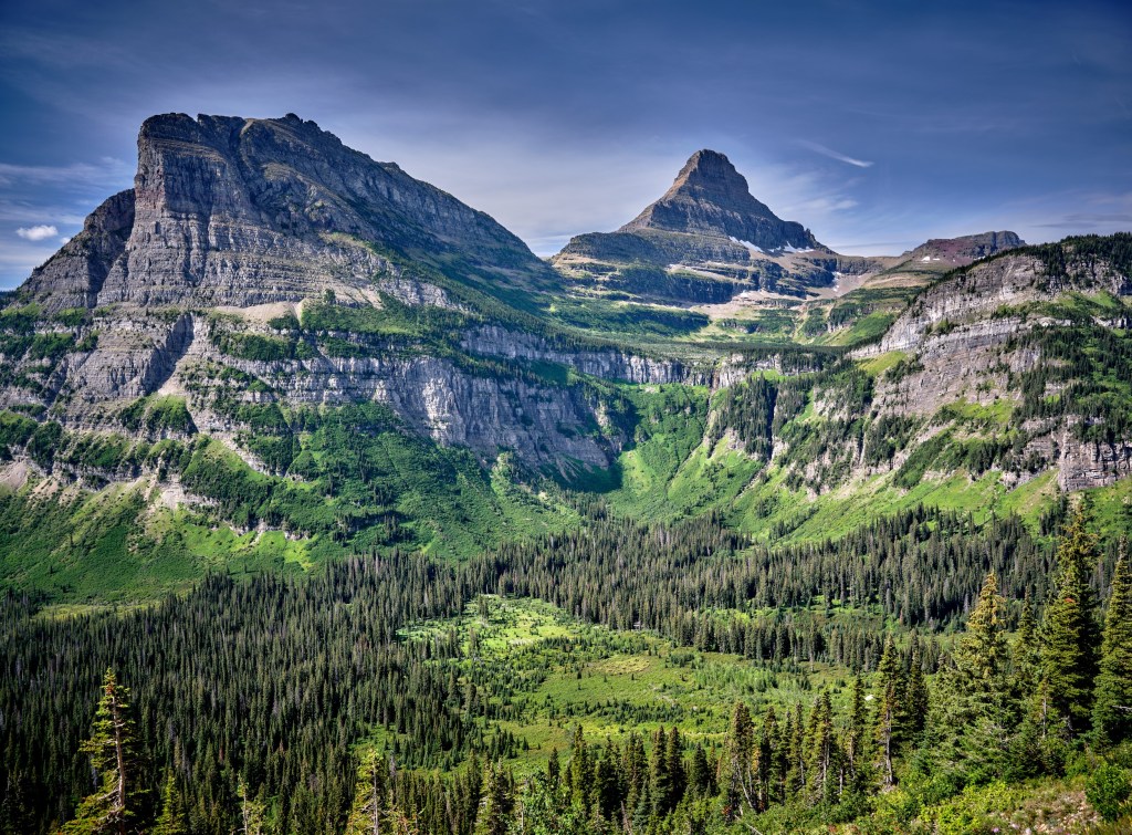 Rugged peaks rise above a lush valley with bright meadow patches and a winding mountain road view.