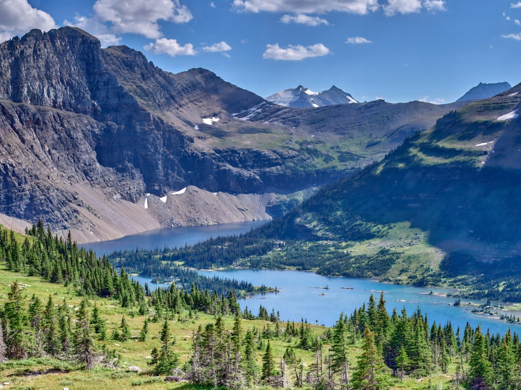 A turquoise lake lies far below rugged cliffs and sunlit mountain ridges viewed from a high trail.