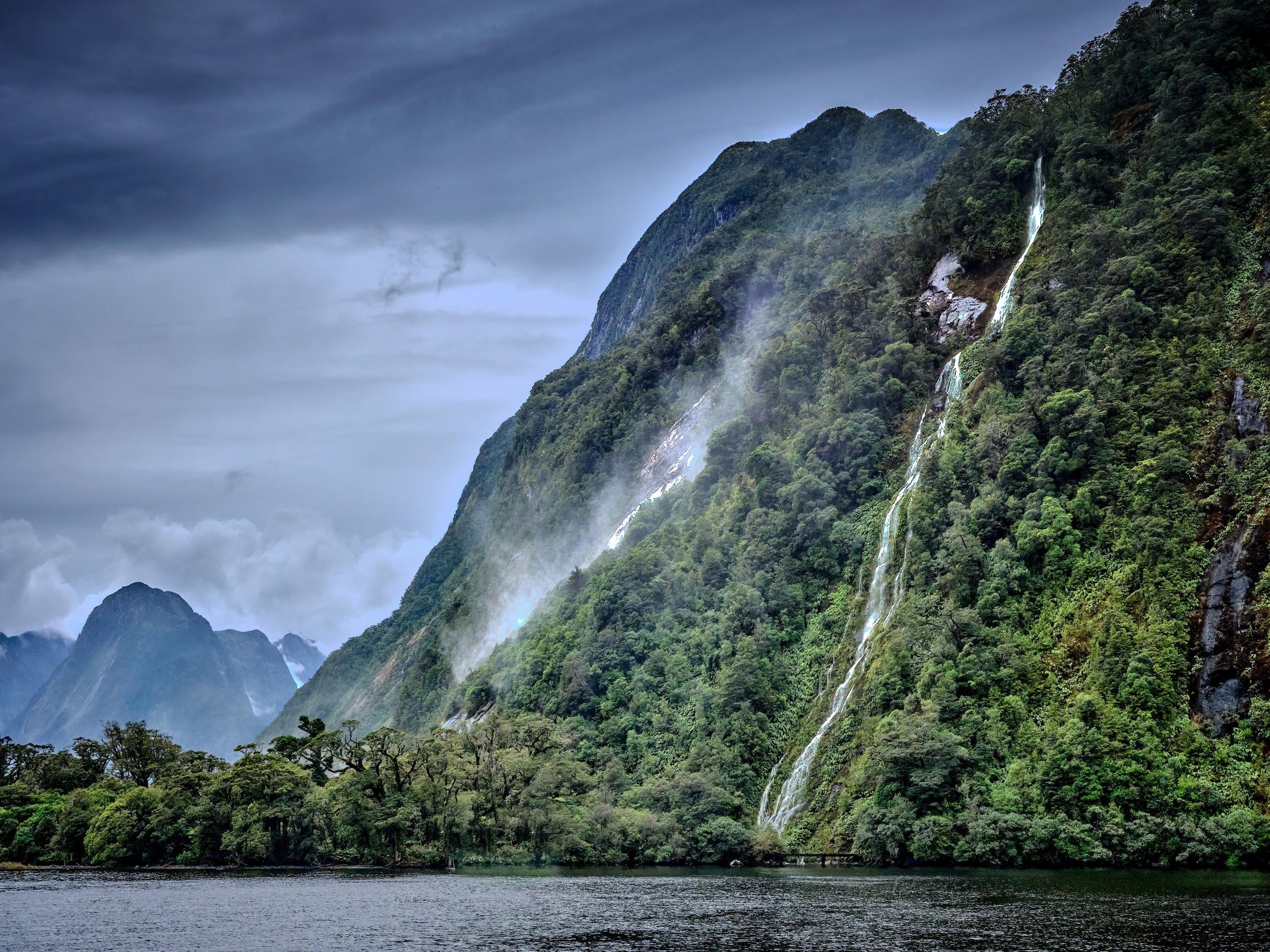 Milford Sound 29 Spray and mist rise beside steep green cliffs near Lady Bowen Falls at Milford Sound.