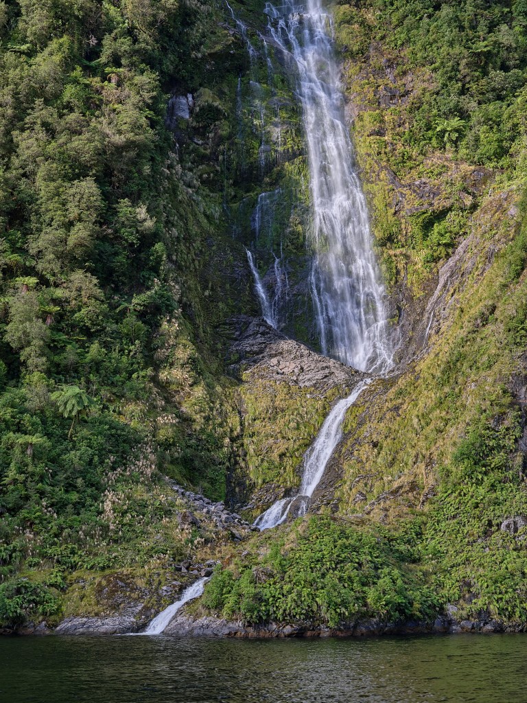 New Zealand 22 A high waterfall splits across dark rock ledges surrounded by dense green vegetation.