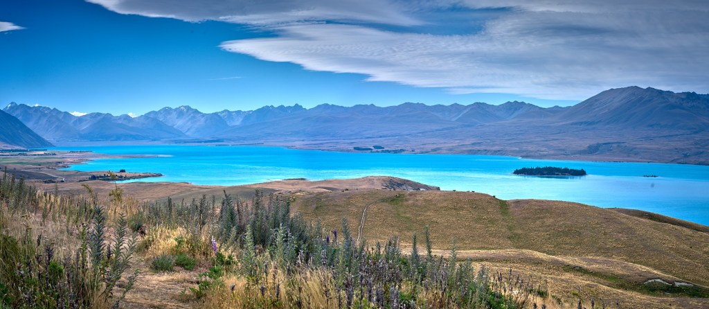 New Zealand 51 A vivid blue glacial lake stretches below golden grasses and distant mountain ranges.