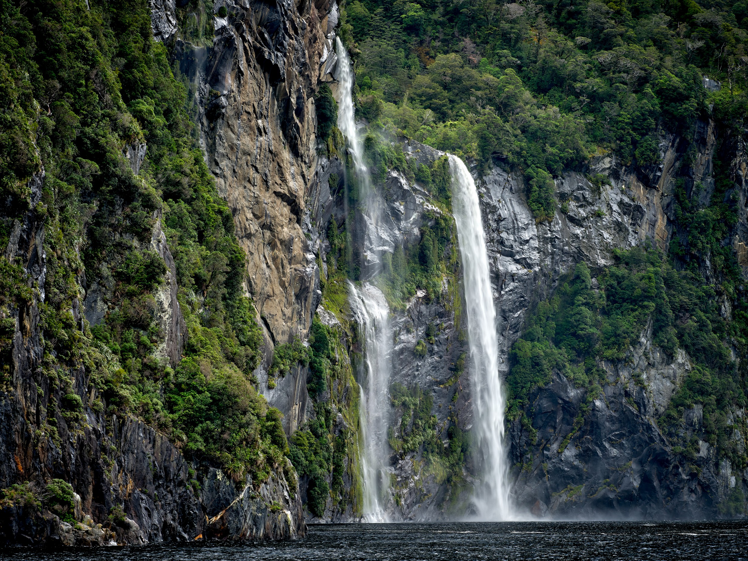 Milford Sound 3 Spray rises where Stirling Falls crashes beside a tour boat along the sheer cliffs of Milford Sound.