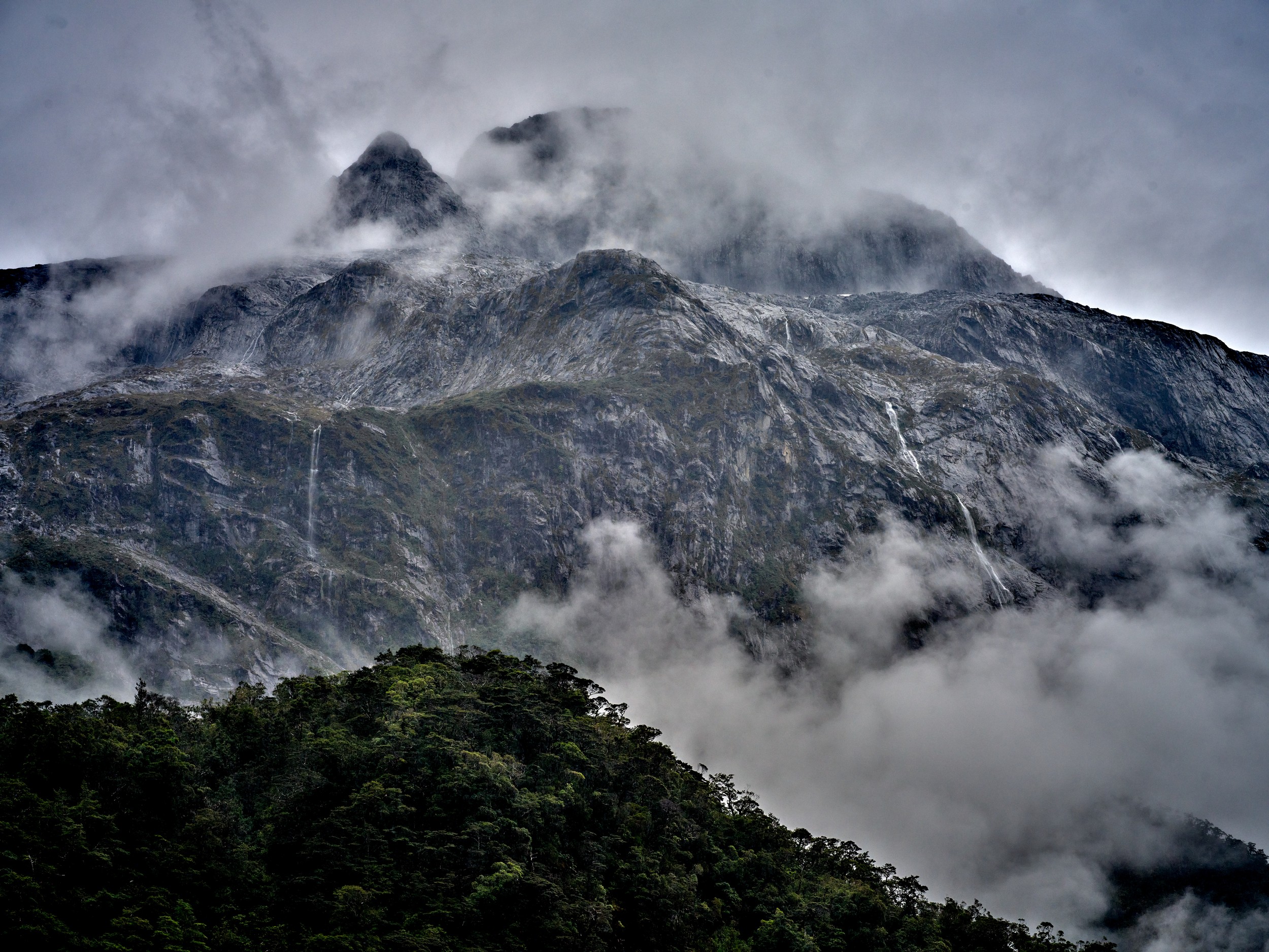 Milford Sound 2 Low storm clouds weave around rocky peaks above dense forest near Milford Sound after rain.