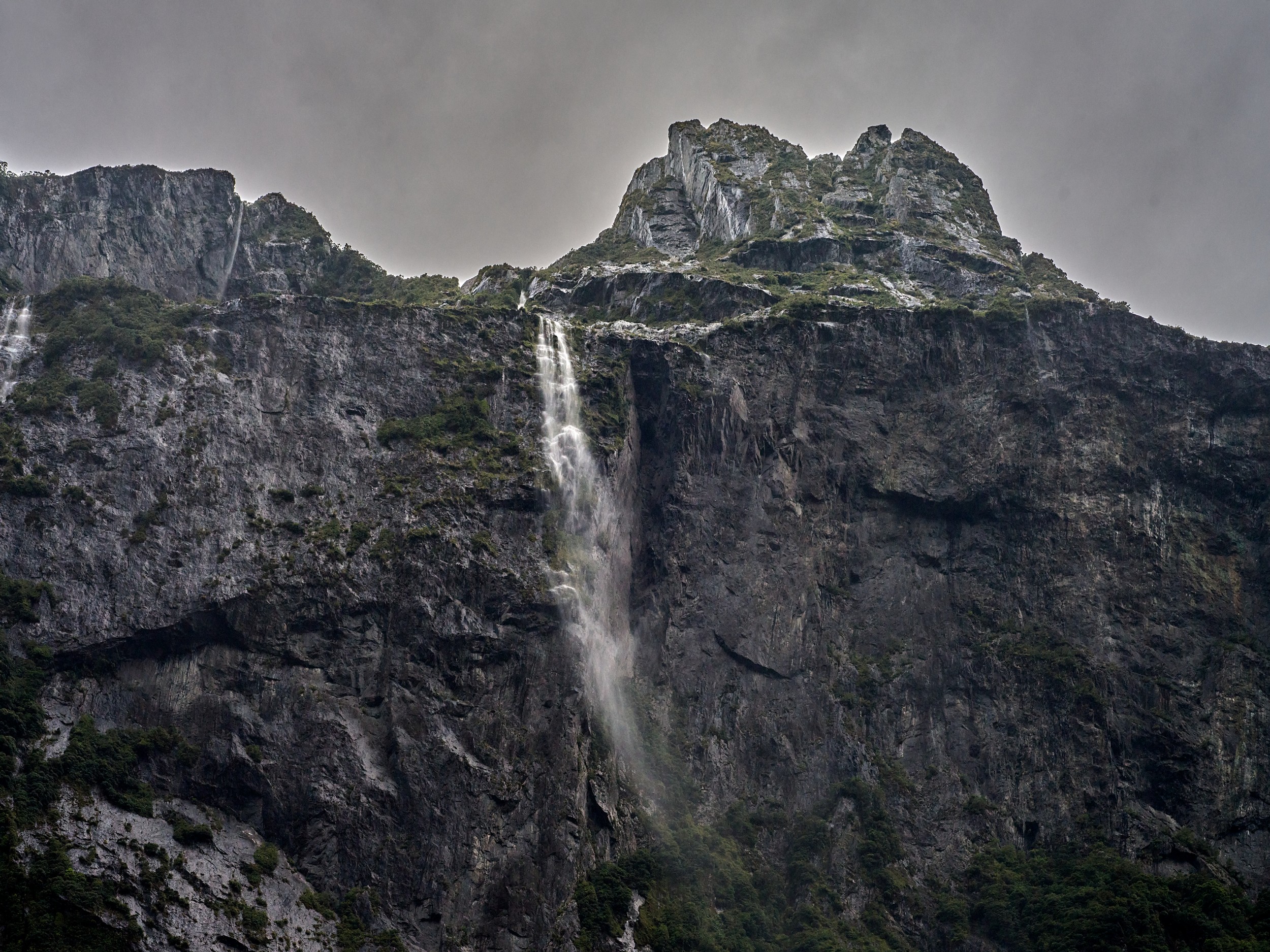 Milford Sound 19 A narrow waterfall cuts down a jagged gray cliff beneath broken cloud cover in Milford Sound.