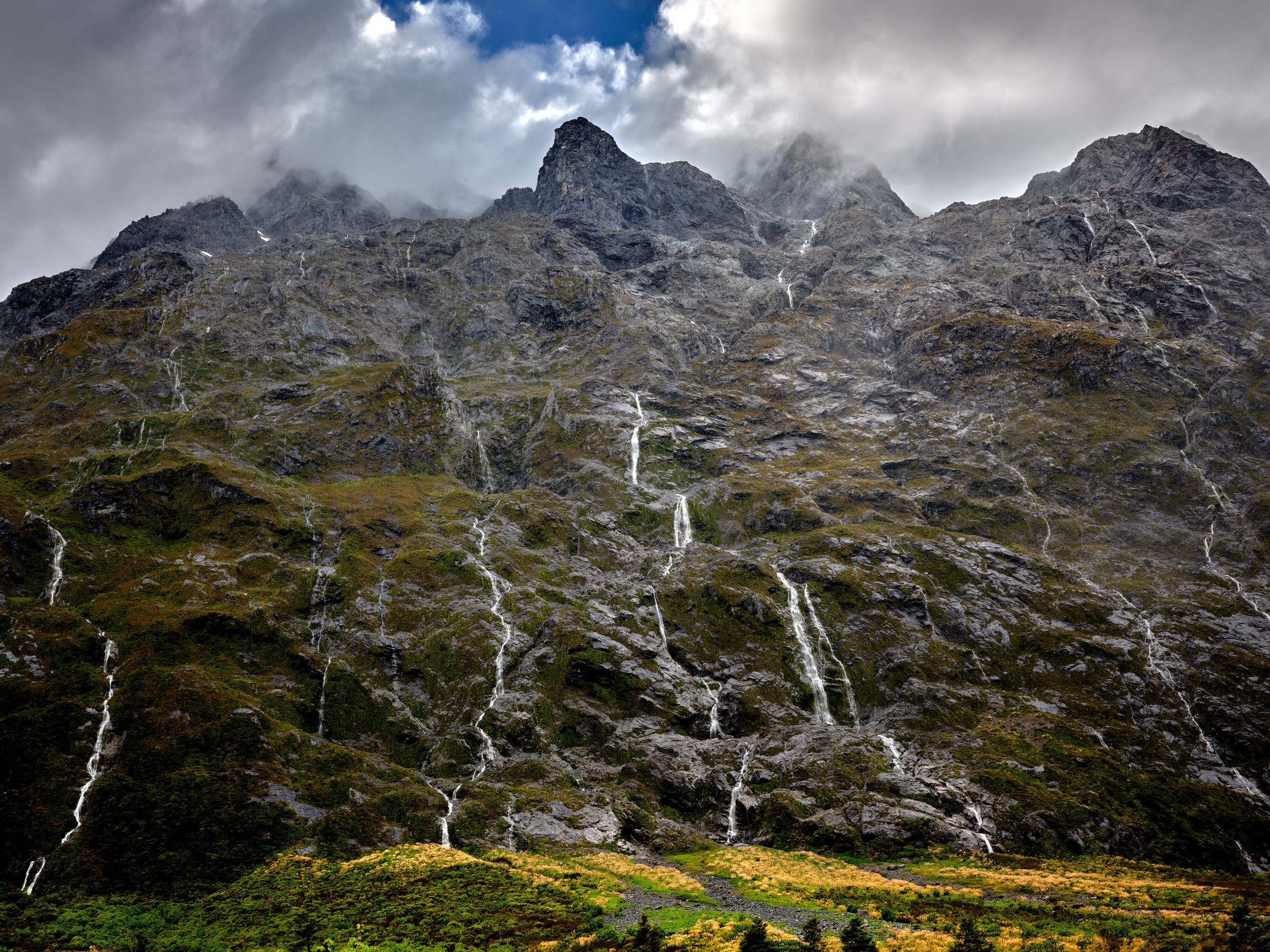 Milford Sound 25 Fresh rain-fed waterfalls stream down a rugged mountain slope beneath broken clouds near Milford Sound.