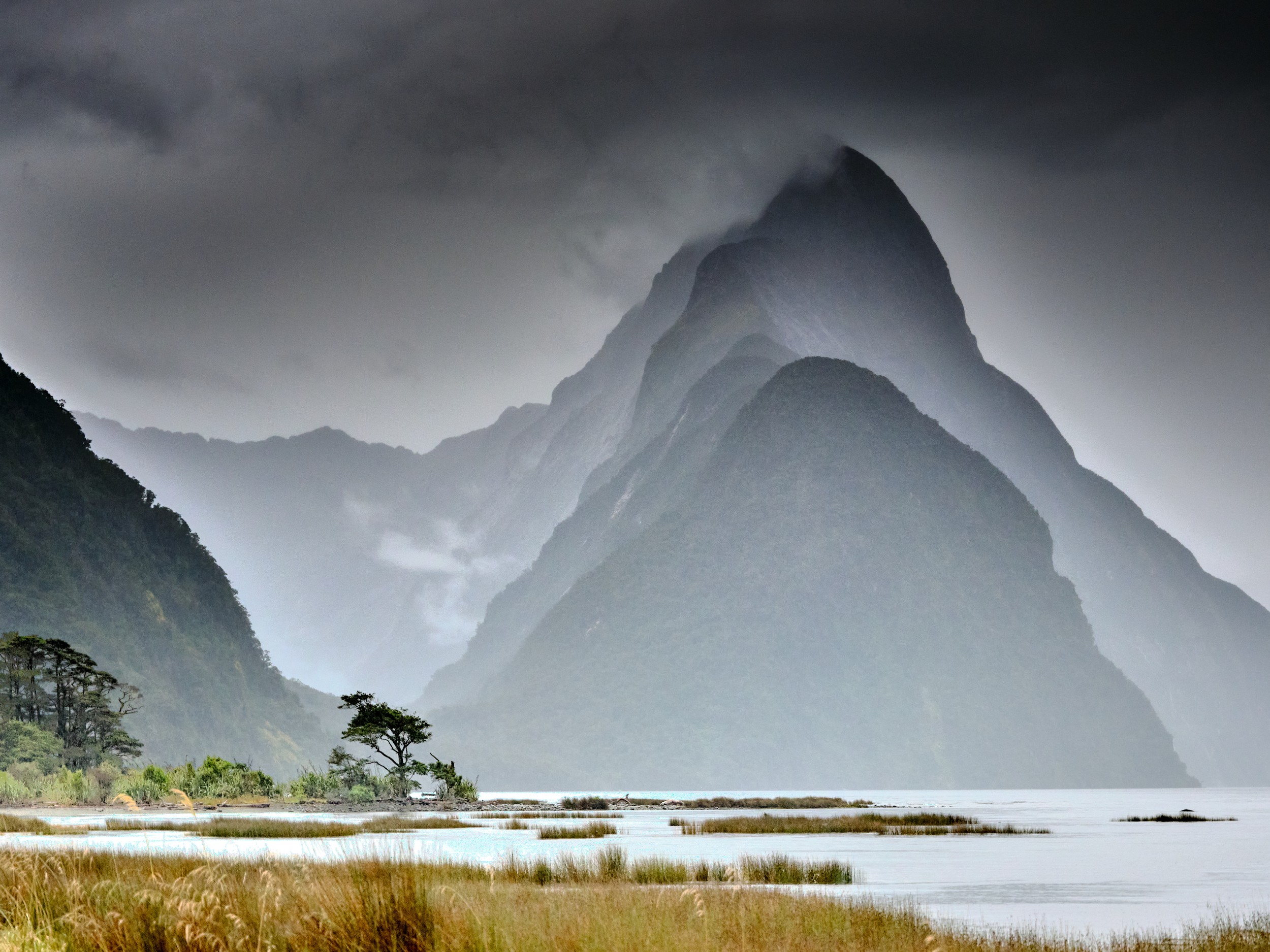 Milford Sound 7 Mitre Peak rises sharply through mist and dark clouds above the shoreline of Milford Sound.