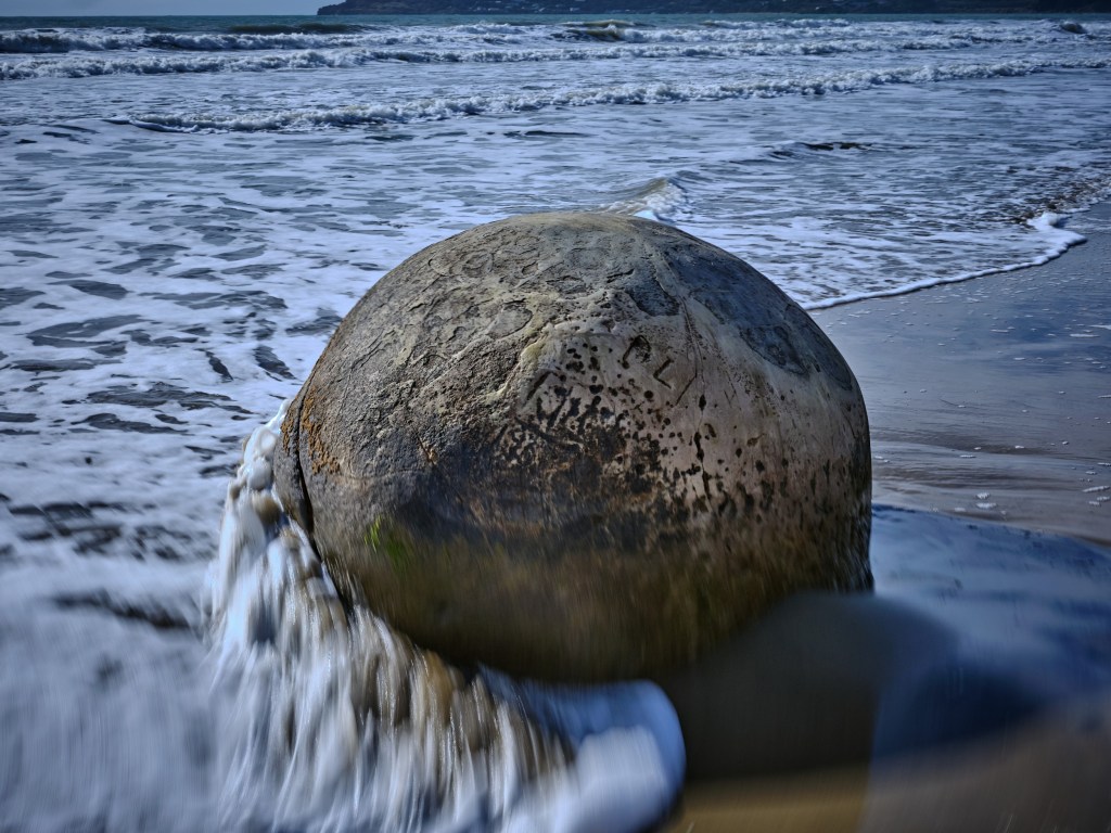 New Zealand 69 A spherical stone sits on wet beach sand while blurred waves rush around it.