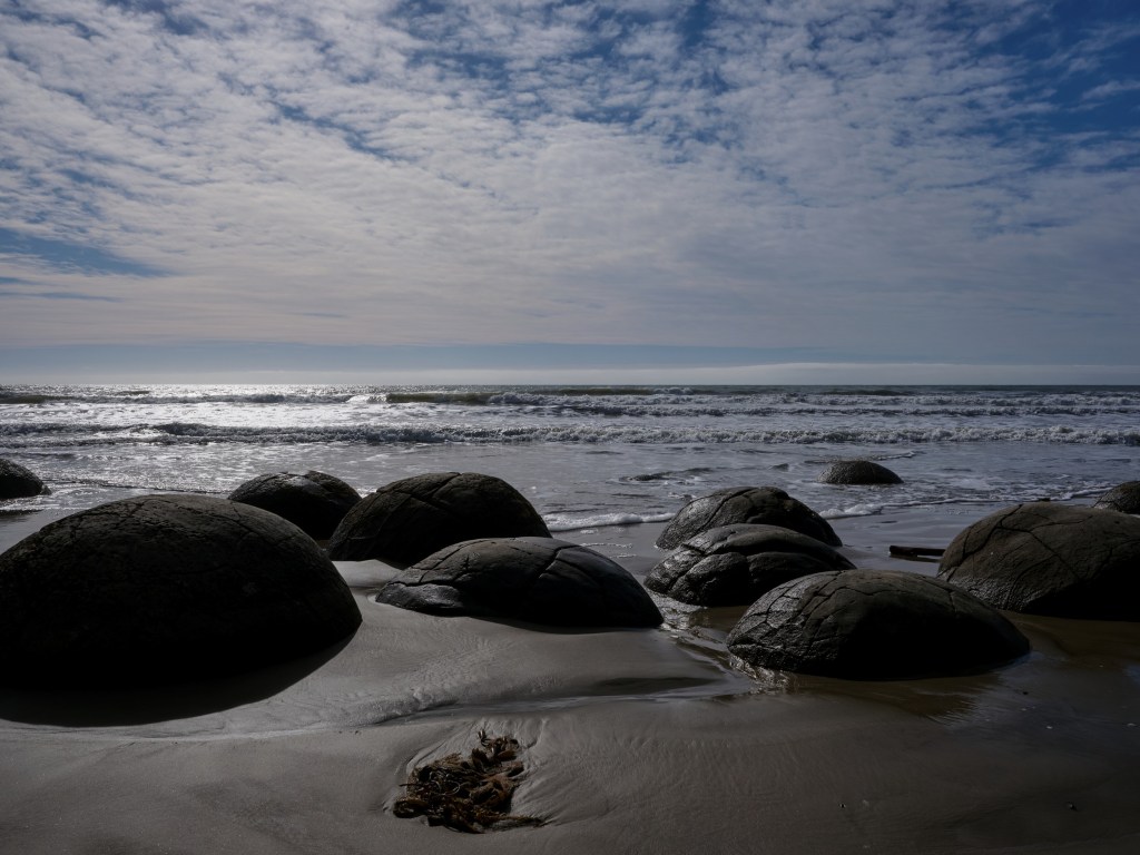 New Zealand 62 Rounded coastal stones rest on dark wet sand beneath patterned clouds and ocean surf.