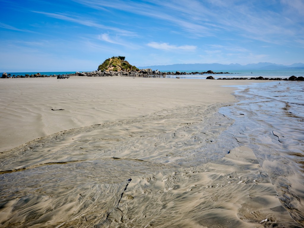 New Zealand 5 Pale sand and shallow water lead toward a rocky islet beneath a clear coastal sky.