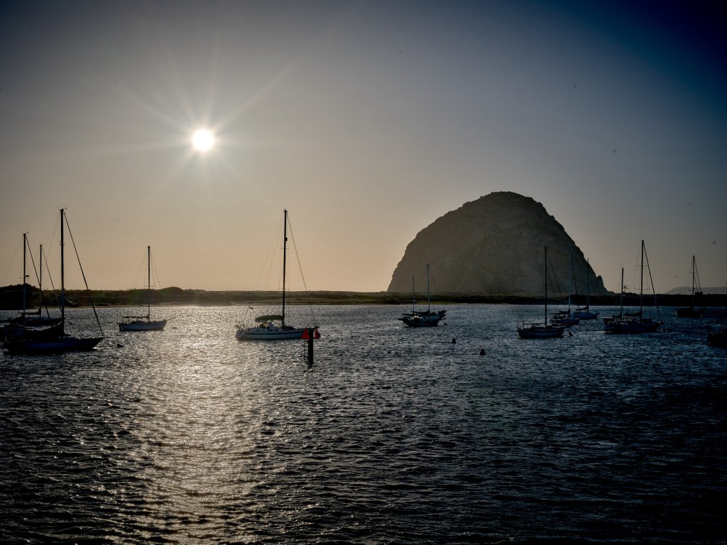 Harbor masts and small boats appear in silhouette against bright water and the volcanic landmark.