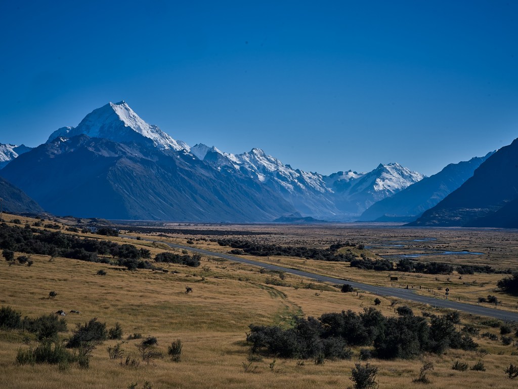 New Zealand 83 A sharp snow capped peak rises beyond open scrubland and distant blue mountain ridges.