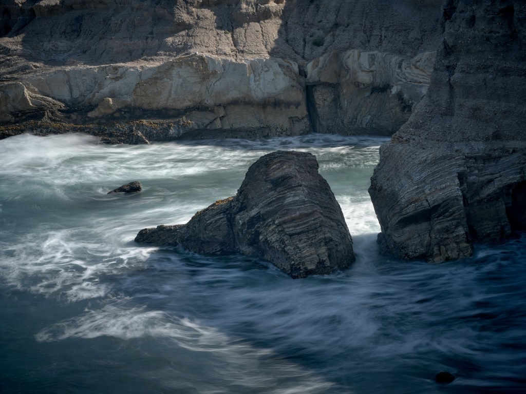 Strong waves push into a cliff-lined pocket with rounded rocks and dramatic coastal shadows.