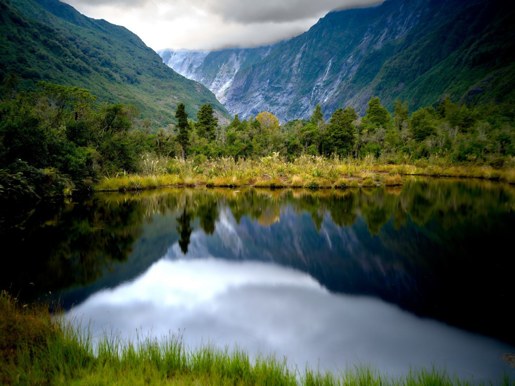 New Zealand 72 A calm pond mirrors trees and clouded mountains near the former foot of Franz Josef Glacier.
