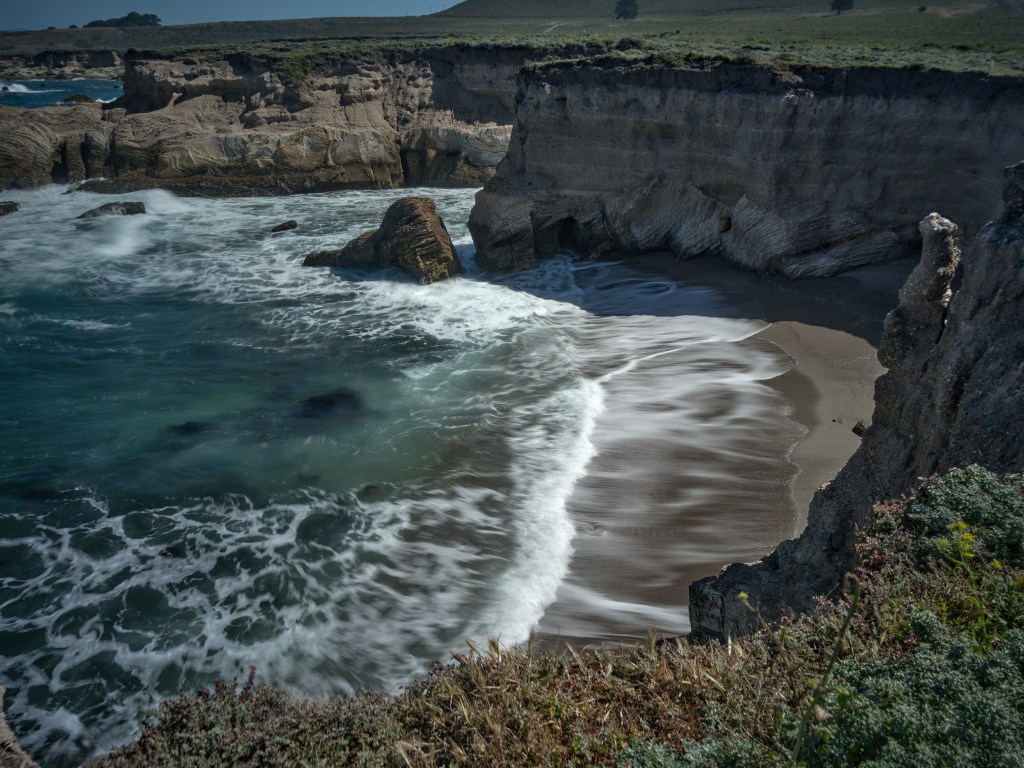 White foam reaches a secluded sandy cove enclosed by eroded walls and rocky headlands.