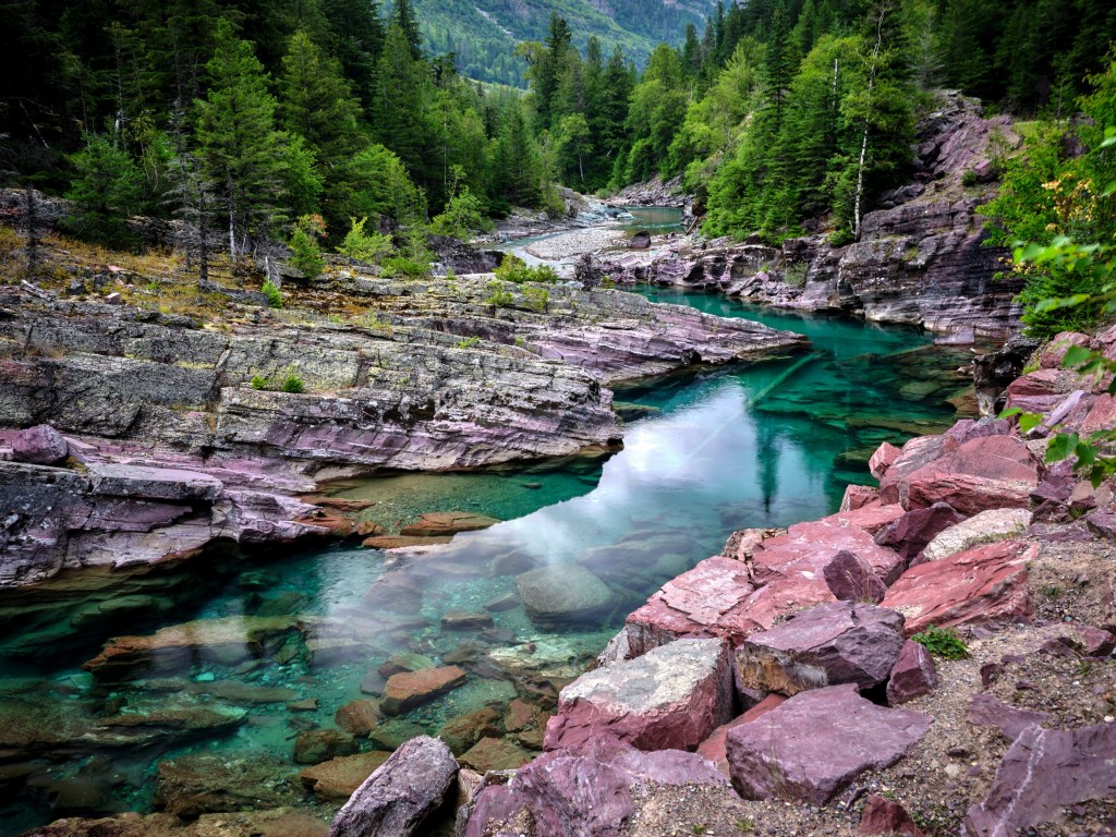Clear green-blue water winds past reddish rocks and evergreen trees under bright mountain light.