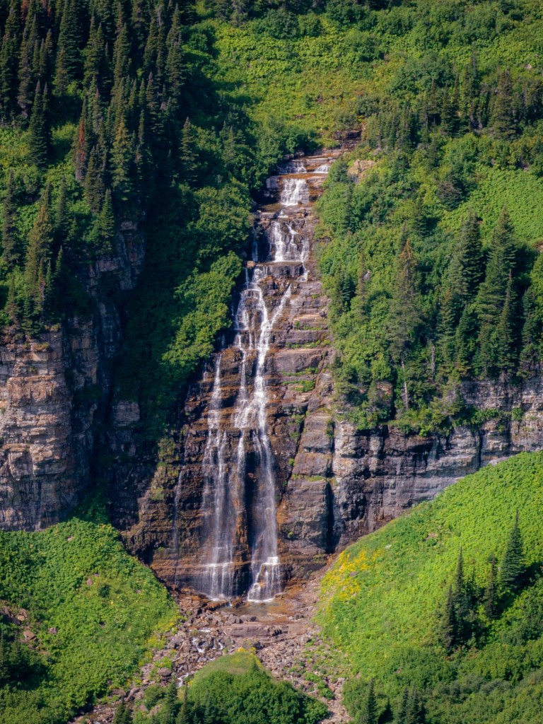 A distant waterfall drops down a steep cliff surrounded by dense green vegetation and forested slopes.