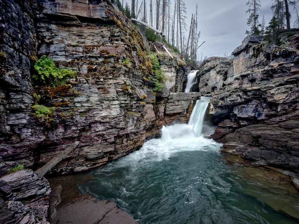 White water spills into a clear turquoise pool enclosed by steep layered stone and forest.