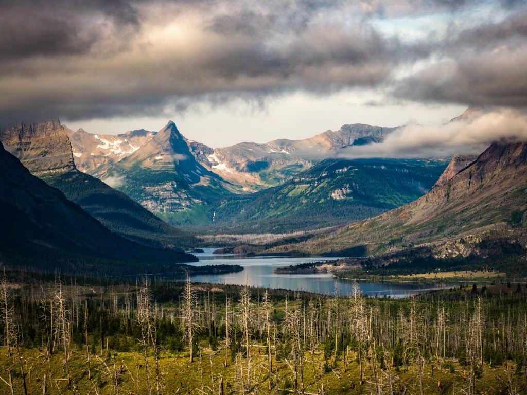 A wide lake sits beneath moody clouds, with mountains and forested slopes receding into the distance.