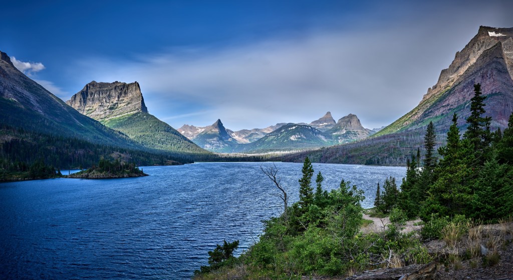 Panoramic view of a deep blue mountain lake bordered by evergreens, cliffs, and distant summits.