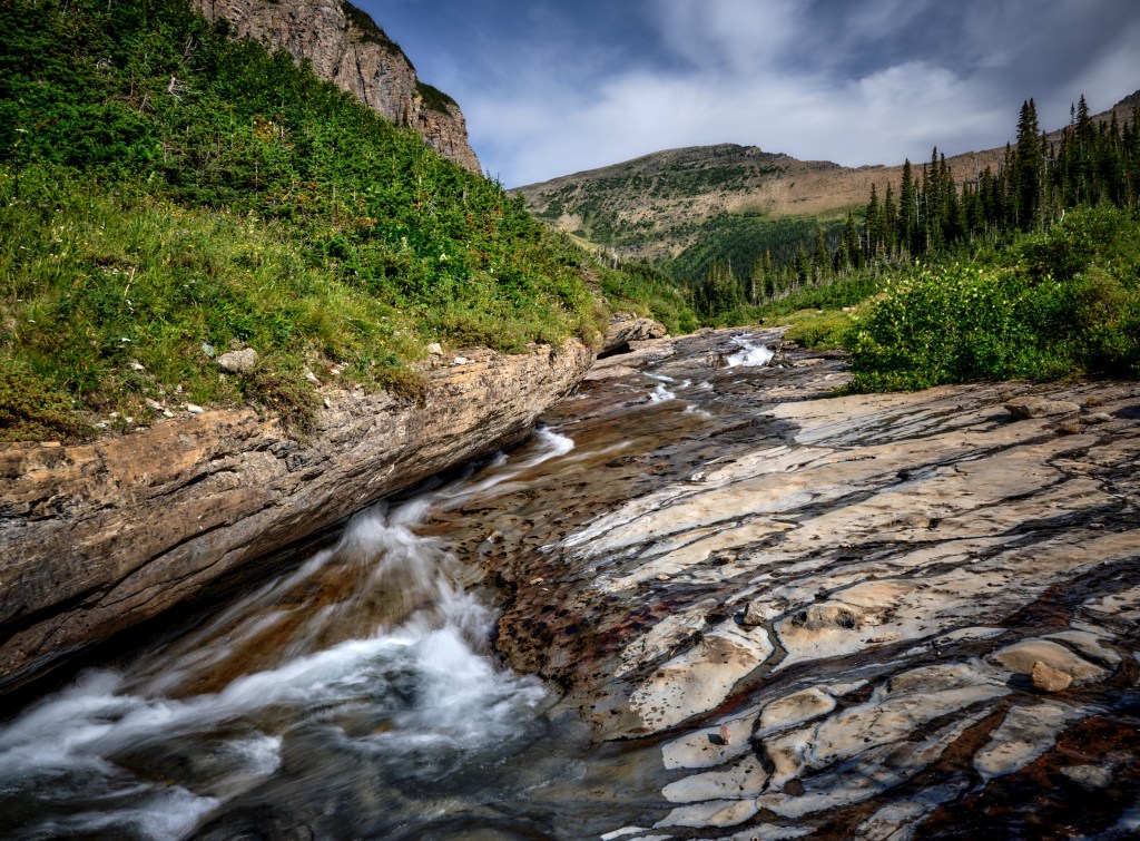 A stream rushes over tilted tan and gray stone slabs beneath low green alpine vegetation.