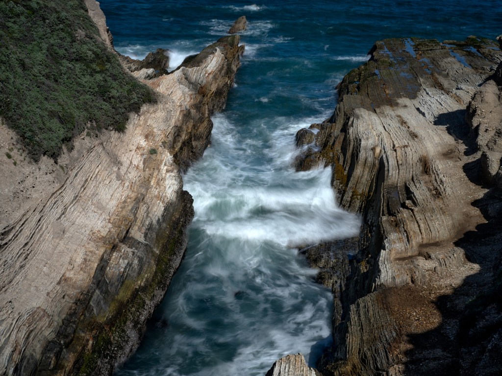 Blue waves squeeze between steep sedimentary walls, throwing whitewater into the channel.