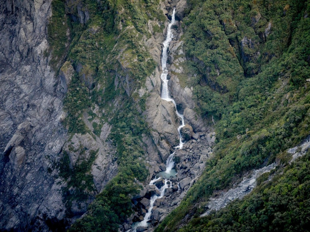New Zealand 7 A tall mountain waterfall threads down green cliff walls toward the glacier landscape below.