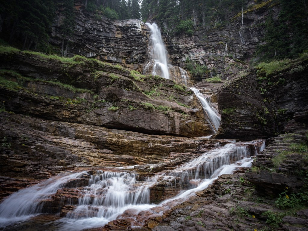 Tiered white cascades descend over dark rock shelves with green plants tucked along the stream banks.