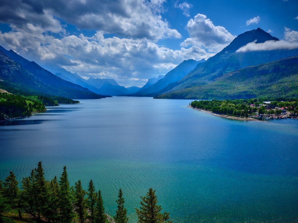 A vivid mountain lake extends into the distance with forested shores and cloud-dappled sky above.