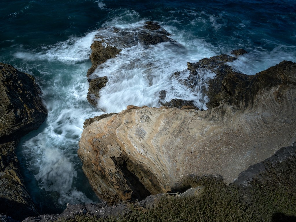 Ocean foam spreads over jagged tan and dark rocks at the base of the bluff.