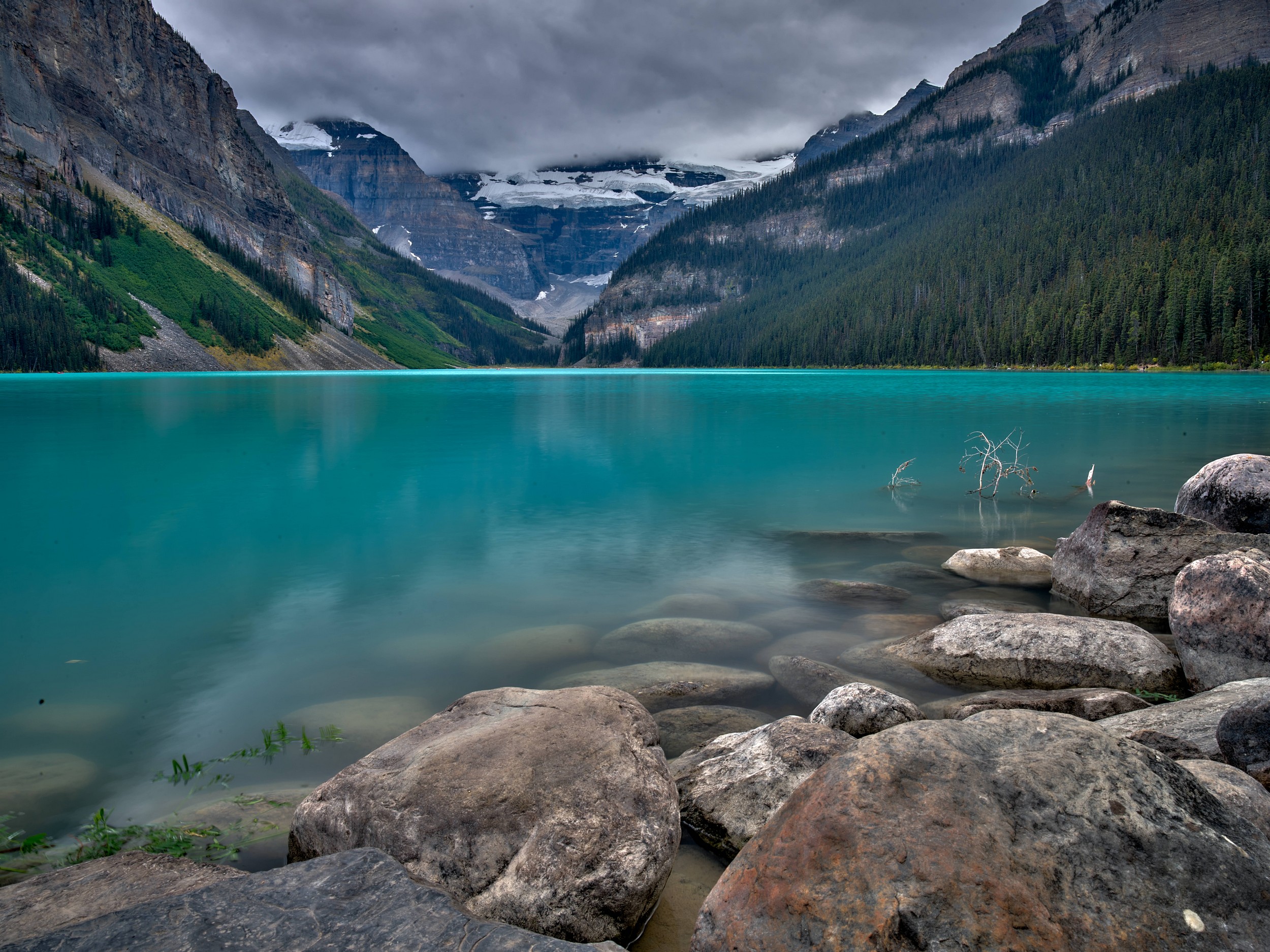 Turquoise Lakes 11 Smooth gray boulders fill the foreground beside vivid lake water, forested mountains, and distant glacier ice.