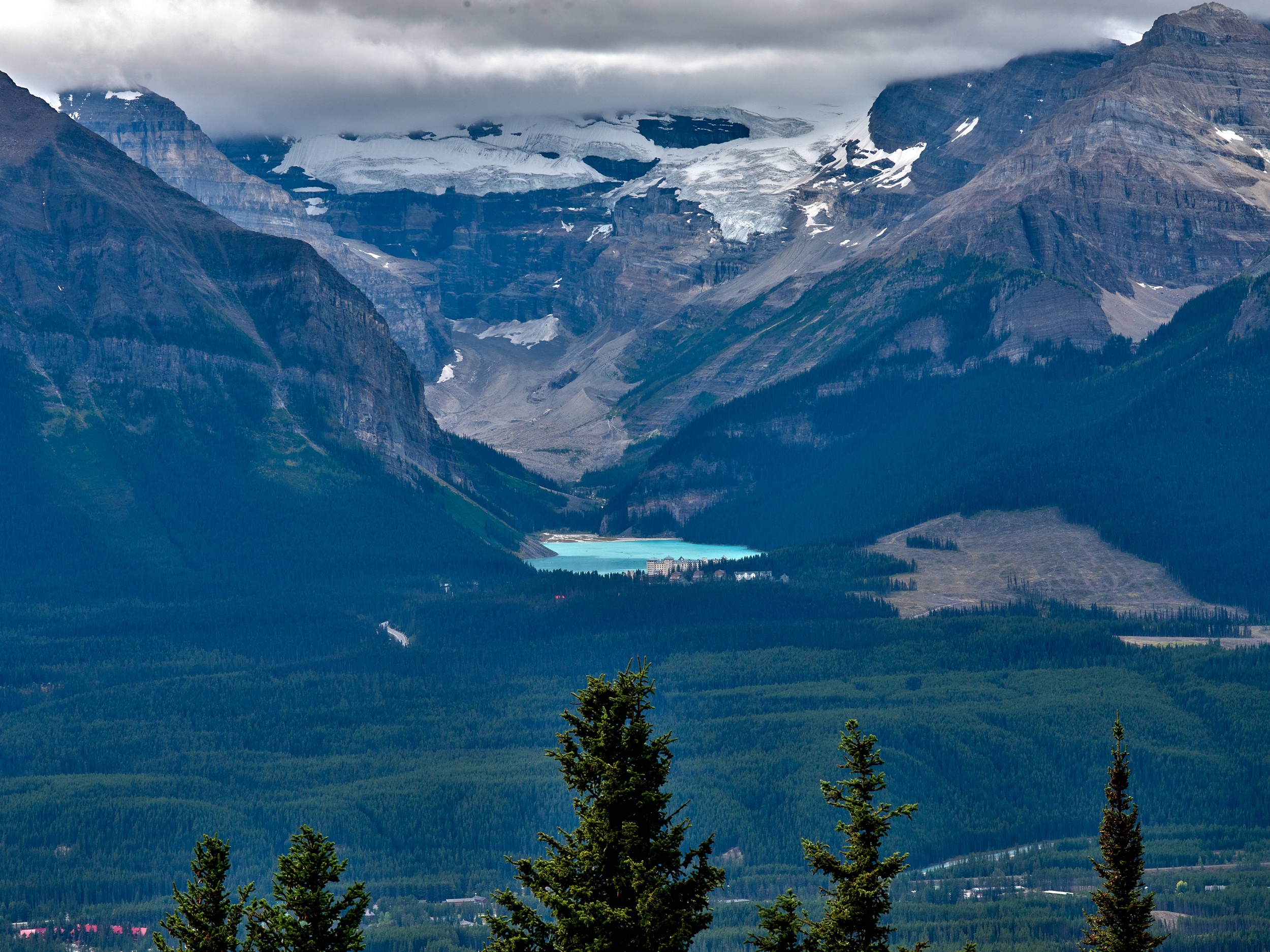 Turquoise Lakes 19 A small turquoise lake lies below pale glacier ice, dark forest, and steep layered cliffs under cloud.