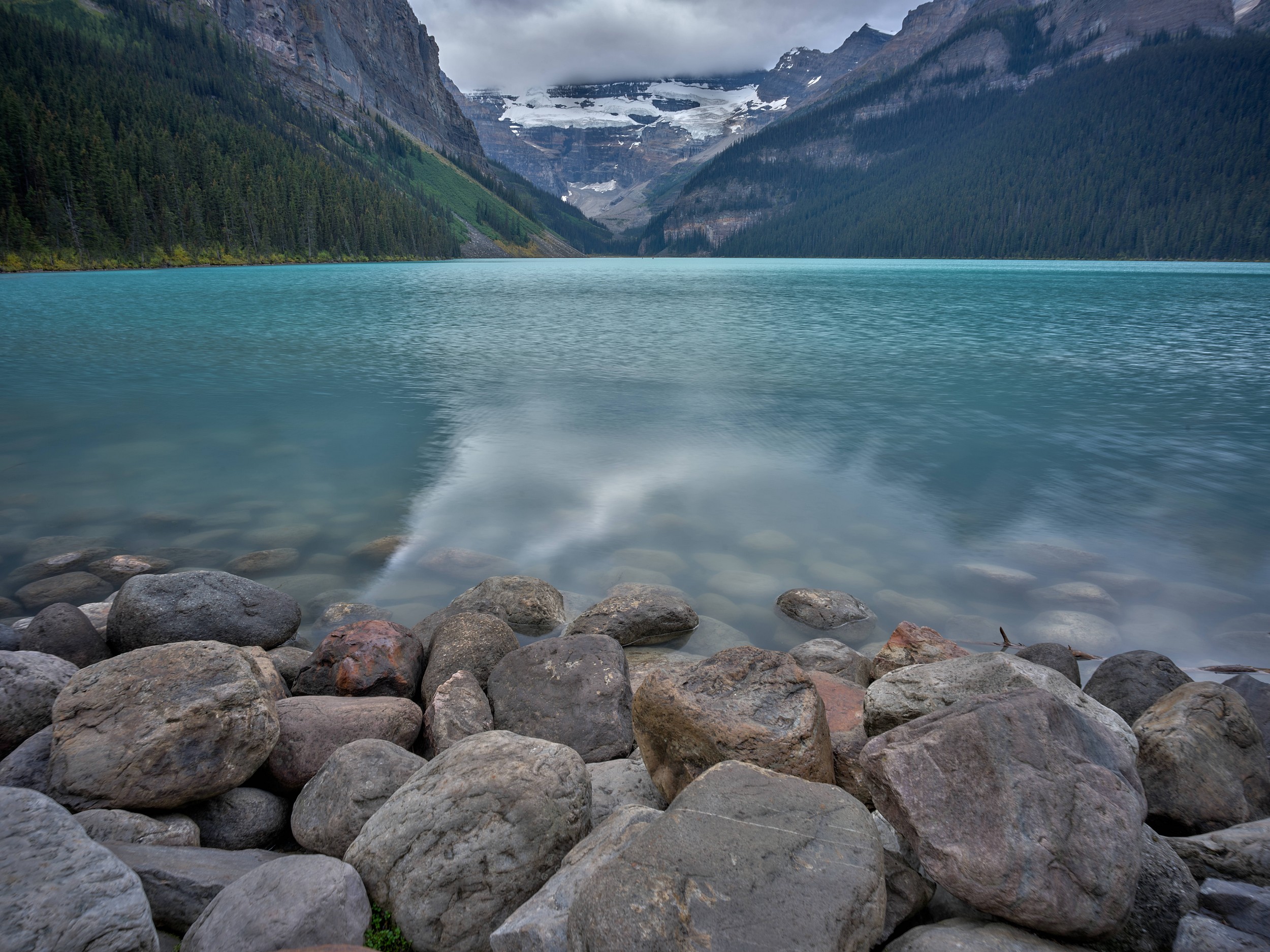 Turquoise Lakes 1 Rounded gray boulders line still turquoise Lake Louise with dark mountains and glacier ice in the distance.