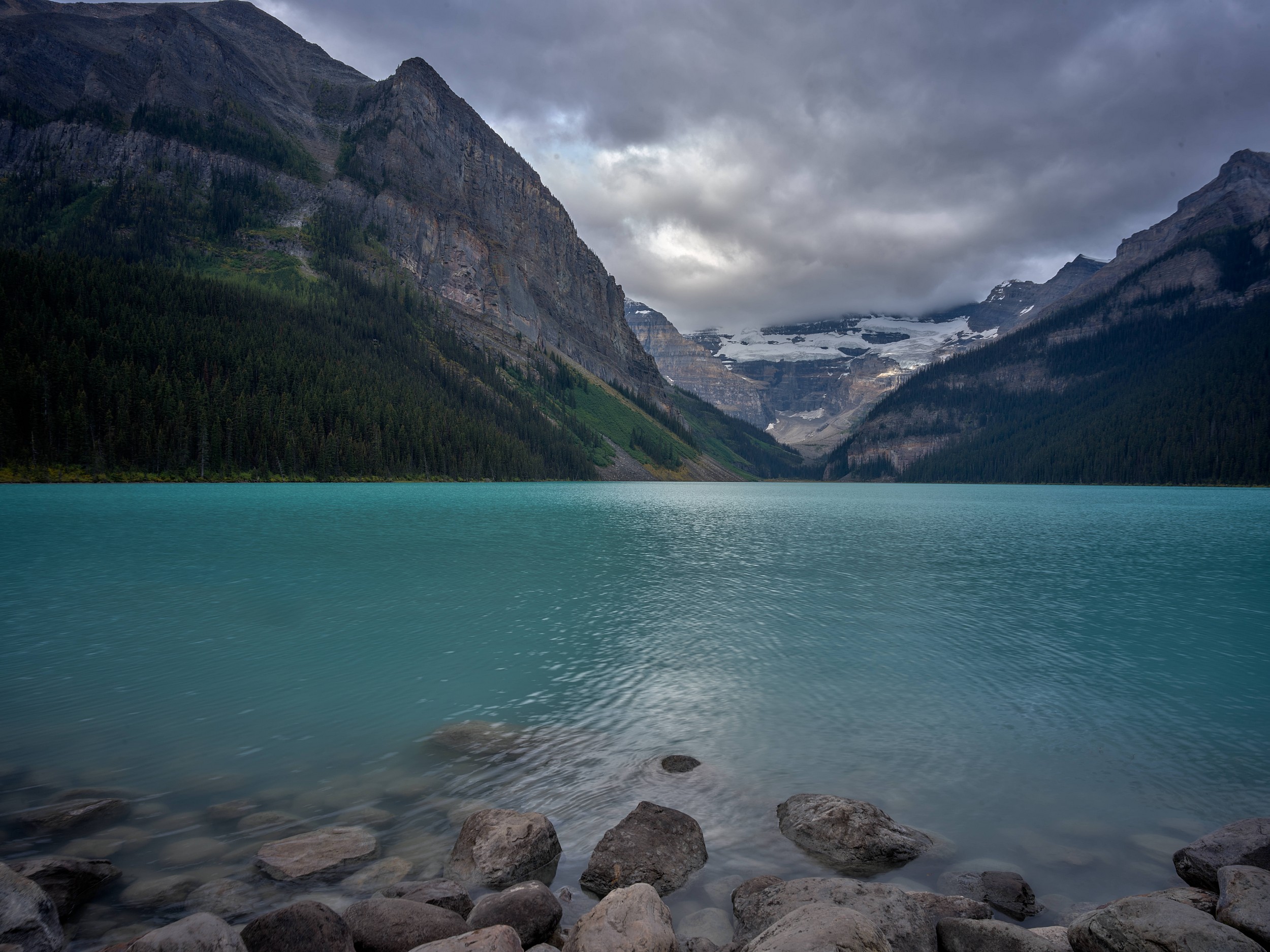 Turquoise Lakes 15 Turquoise water stretches between dark mountain walls toward glacier-covered peaks under overcast skies.