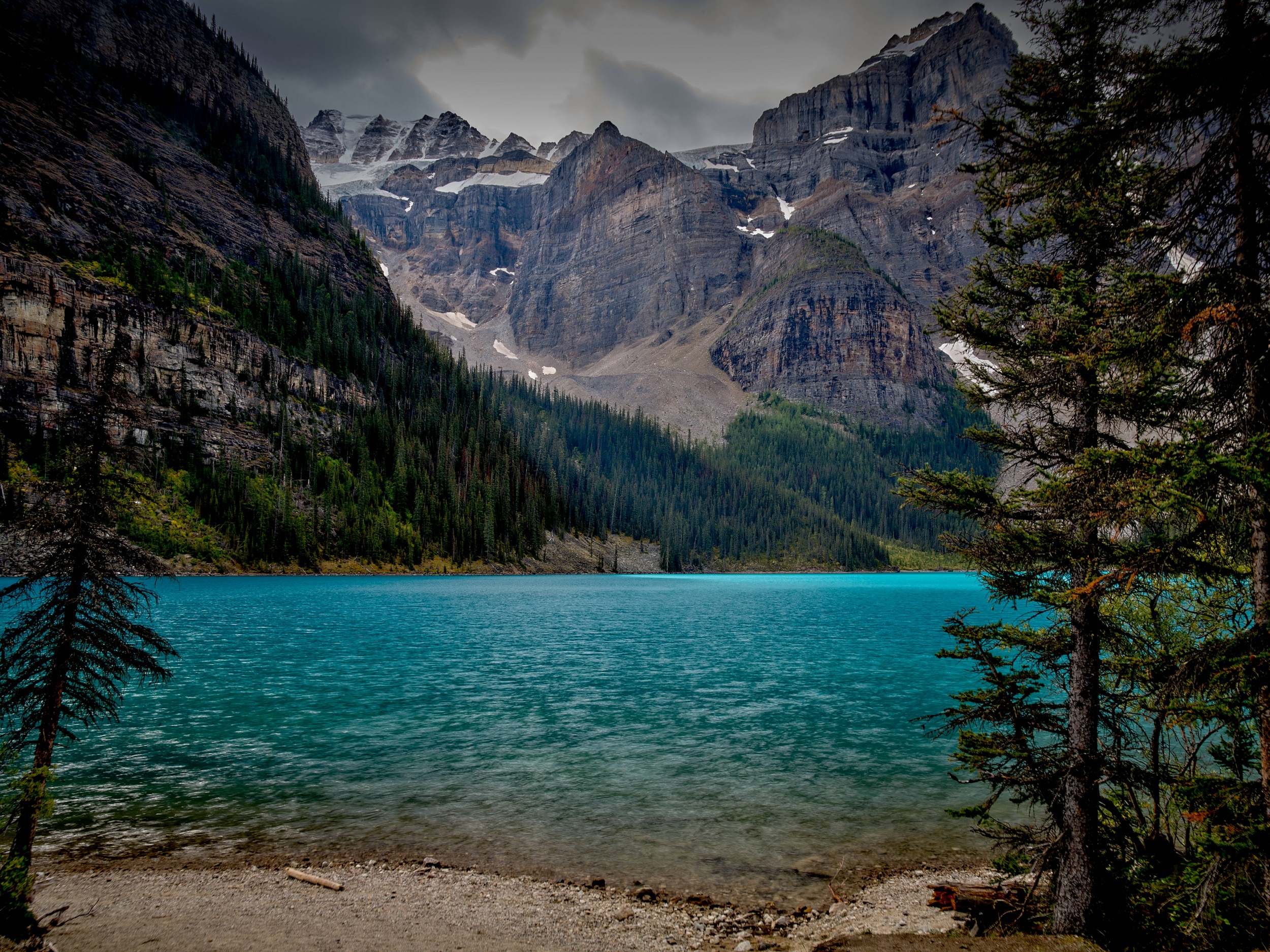 Turquoise Lakes 3 Turquoise Moraine Lake and jagged peaks appear between dark conifers beneath an overcast mountain sky.