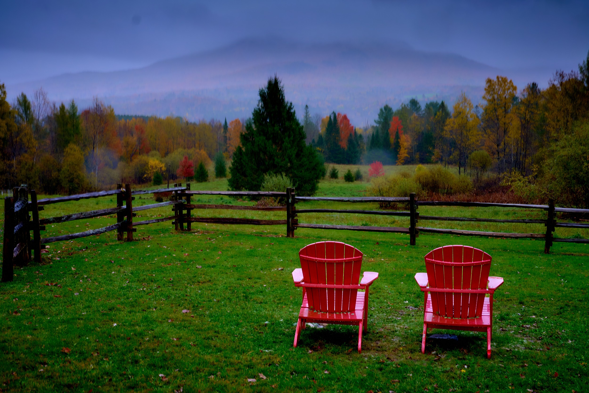 Vermont and Quebec 20 Pair of bright red Adirondack chairs sits beside a dark fence facing misty fall-covered hills.