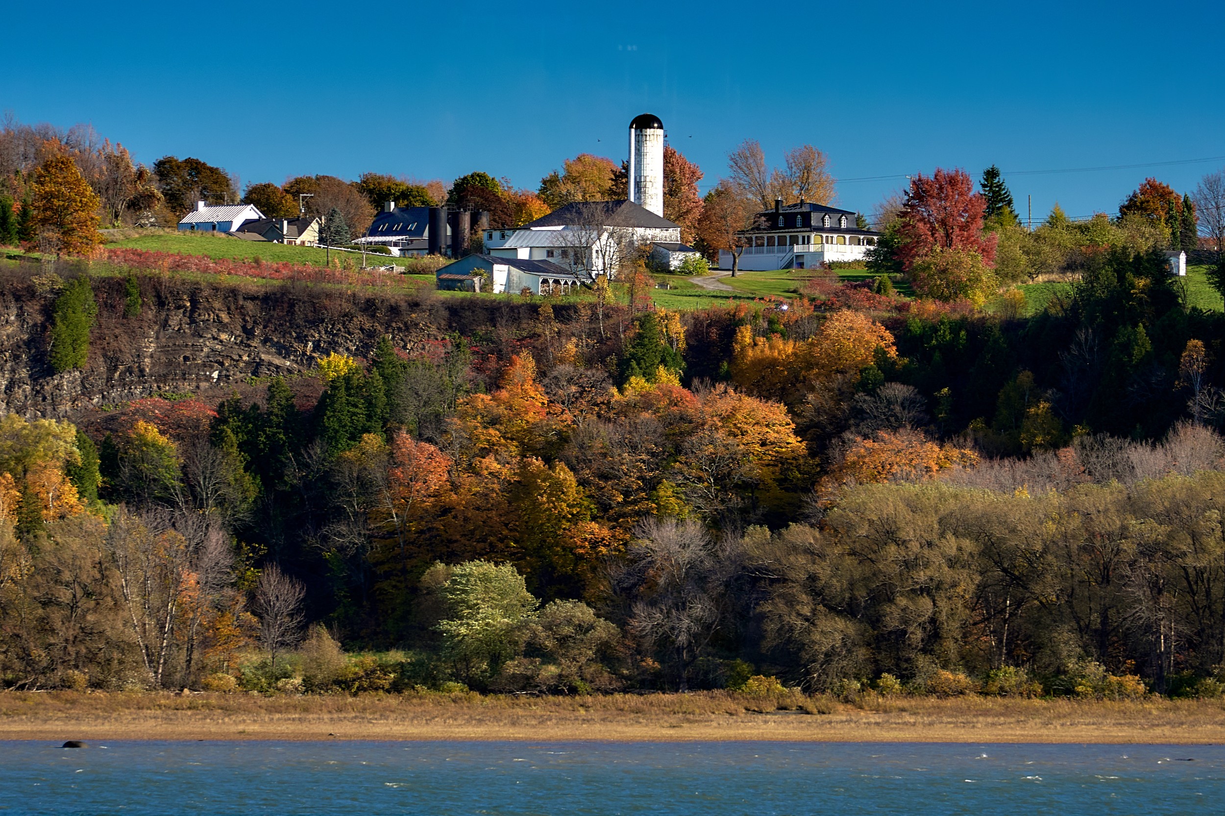 Vermont and Quebec 27 White agricultural buildings with a tall silo sit beyond a narrow shoreline below a patchwork of autumn-colored trees.