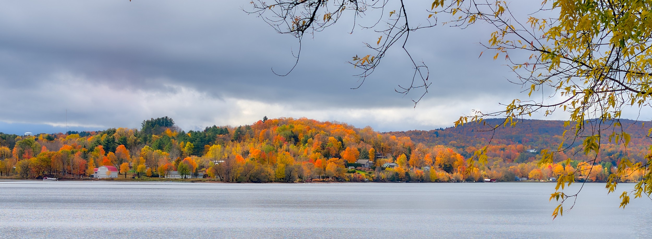 Vermont and Quebec 22 Gray clouds press over calm lake water and a distant band of yellow and orange trees near Newport.