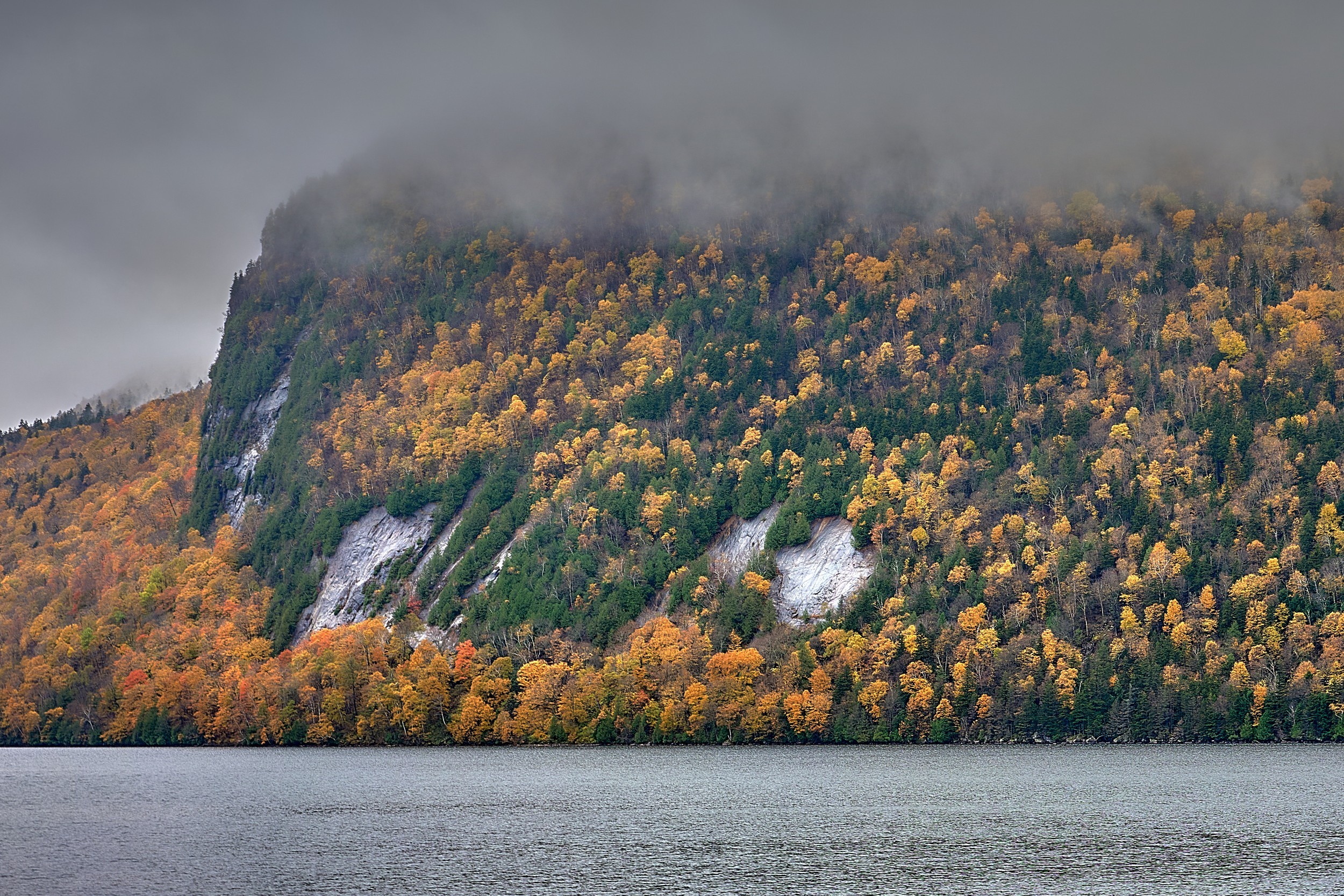 Vermont and Quebec 45 Mist hides the top of a sheer lakeside mountain covered in orange and green forest at Lake Willoughby.
